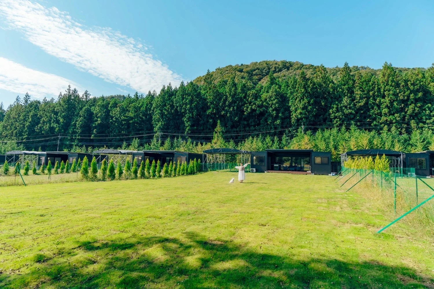 広々とした芝生が広がる敷地にモダンな宿泊施設が並び、一人の女性と白い犬がくつろいでいる